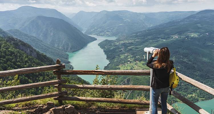 Traveler at a wooden lookout gazes over a winding turquoise river through dramatic mountain valleys.