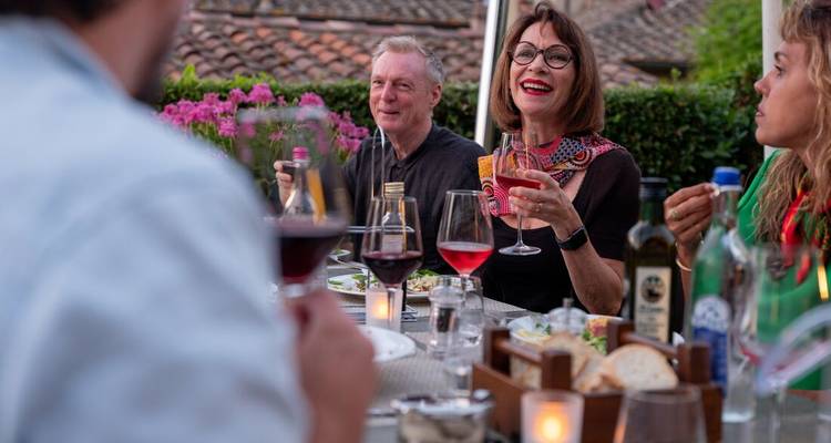 Des amis partagent du vin et un dîner à une table extérieure entourée de fleurs en soirée.