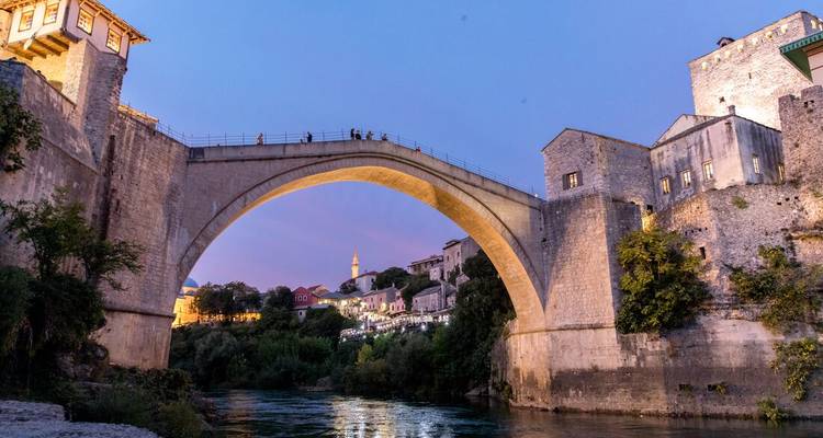 Evening lights glow on Mostar’s Stari Most bridge arching over the Neretva River at twilight.
