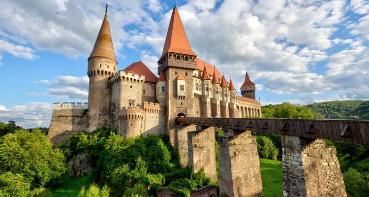 Majestic Corvin Castle with red spires and stone bridge set against a blue sky and fluffy clouds.