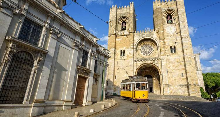 Le tramway historique jaune tourne au coin de la façade en pierre de la Cathédrale de Lisbonne sous un ciel bleu éclatant.
