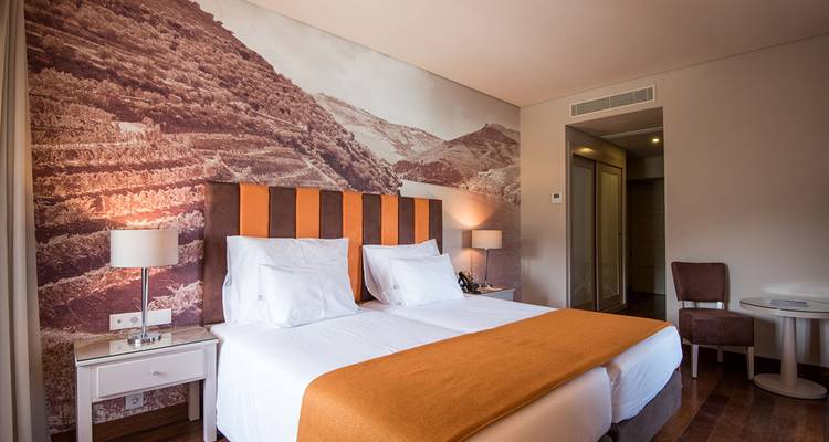 Chambre de boutique hôtel bien éclairée avec deux lits jumeaux impeccablement faits, un bureau et une grande fresque murale à thème viticole derrière la tête de lit.