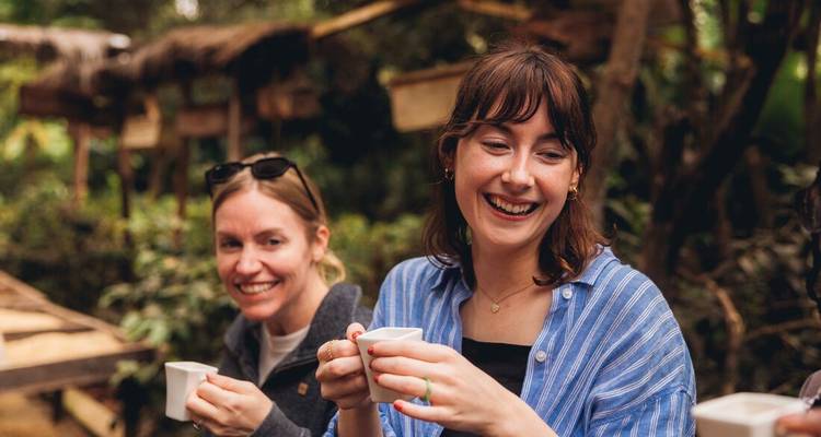Two women smile over cups of fresh coffee at a rustic plantation setting.