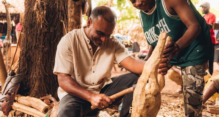Artisan demonstrates wood carving techniques to a traveller in a village setting.