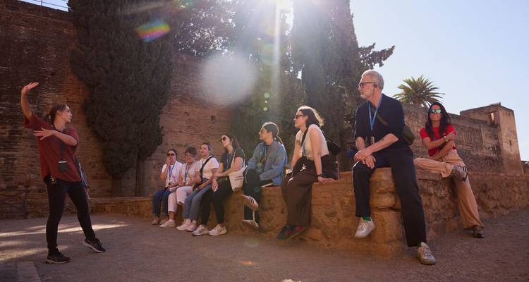 Un guide fait des gestes pendant qu'un groupe de visite assis l'écoute dans une cour ensoleillée à côté de murs anciens.