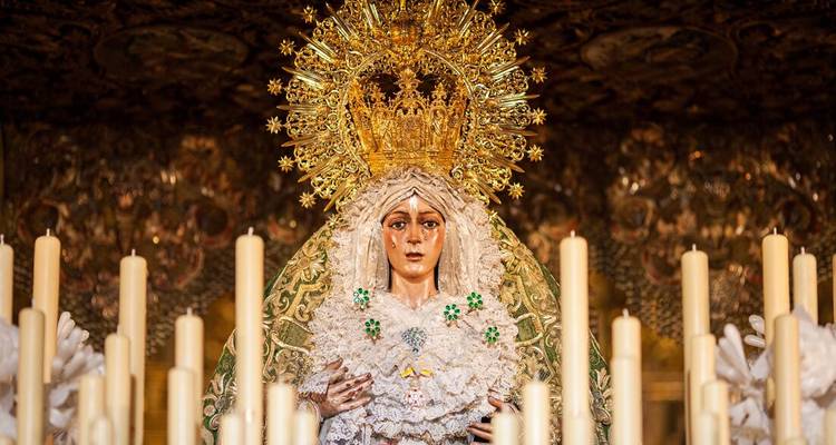 Statue dorée ornée de la Vierge Marie entourée de rangées de grands cierges dans une chapelle faiblement éclairée.