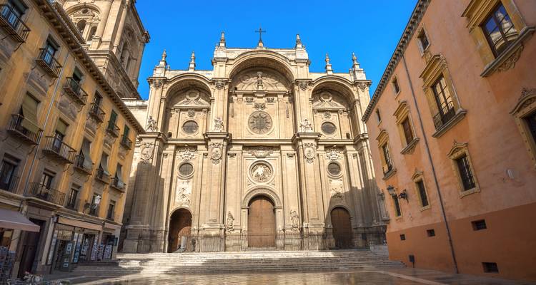 La fachada de piedra de la Catedral de Granada se alza sobre una plaza tranquila y bañada por el sol, rodeada de edificios históricos.