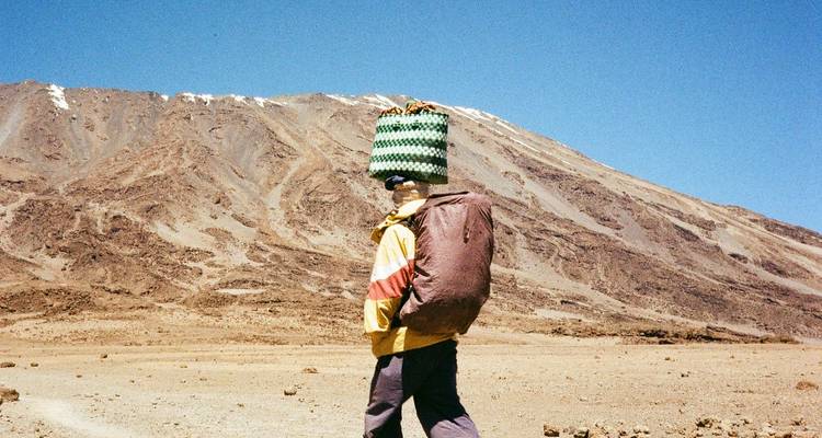 Mountain porter carrying gear basket on head beneath the rocky slopes of Kilimanjaro.