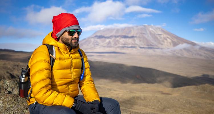 A lone hiker in a yellow puffer jacket and red beanie sits on a rocky ledge with Mount Kilimanjaro dominating the clear, sunny horizon behind him.