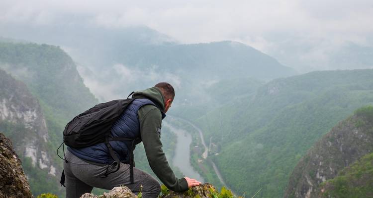 Excursionista masculino con mochila agachado en el borde de un acantilado con vistas a un valle de río brumoso.