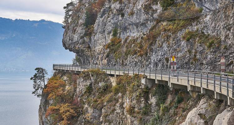 Carretera estrecha construida a lo largo de un acantilado rocoso con vista a un lago tranquilo en un día de otoño.