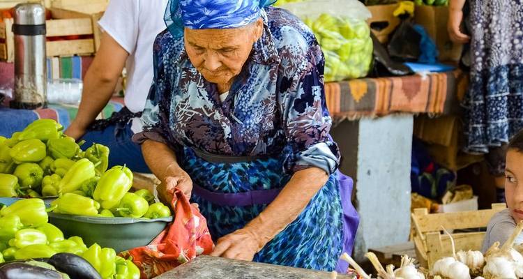 An elderly woman in a blue headscarf sorts green peppers at a bustling market stall.