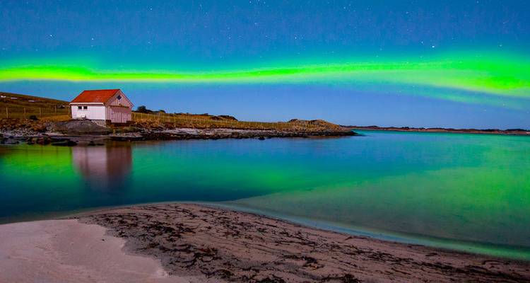 Une aurore d'un vert éclatant s'arque au-dessus d'une baie côtière tranquille avec une cabane solitaire aux toits rouges la nuit.