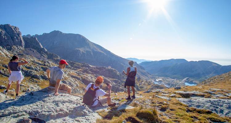 Wandelaars maken een pauze op door de zon beschenen rotsen met uitzicht op een afgelegen alpendorp en een glinsterend meer.