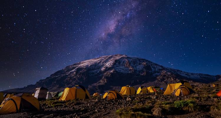 Ciel étoilé et Voie lactée s'arquant au-dessus du Mont Kilimandjaro aux sommets enneigés avec des tentes jaunes au premier plan.
