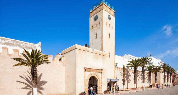 Une tour-horloge de couleur sable s'élève au-dessus des remparts historiques d'Essaouira, encadrée par des palmiers sur un ciel bleu éclatant.