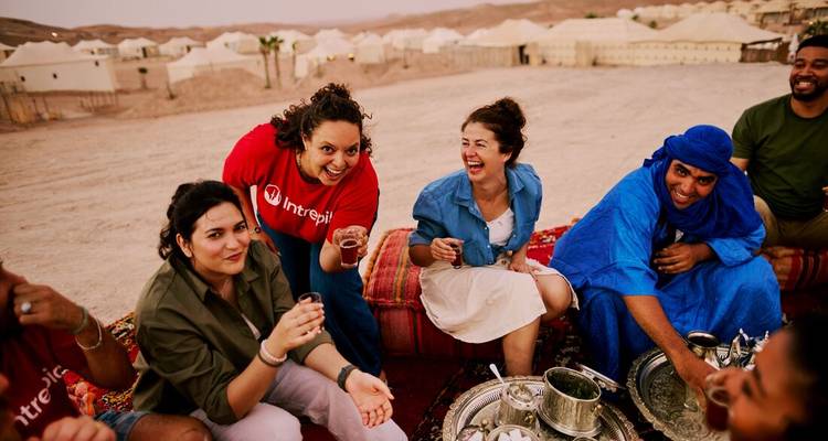 Les voyageurs partagent du thé à la menthe sur des tapis colorés dans un camp désertique avec des dunes dorées au-delà.