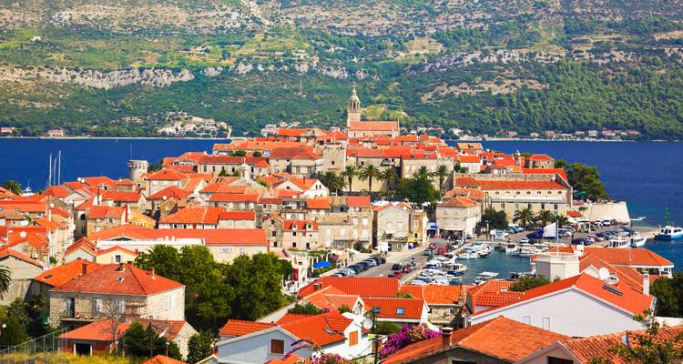 Charming medieval town of Korčula with red-tiled roofs nestled on a peninsula surrounded by deep blue sea and green hills.