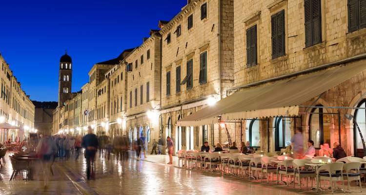 Nachtszene der Stradun mit bewegungsunschärfen Fußgängern und warm beleuchteten Cafés, die sich auf dem nassen Steinpflaster in Dubrovnik spiegeln.