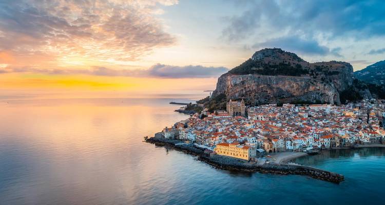 Vista aérea al atardecer de un pueblo costero con casas de techo naranja agrupadas debajo de un promontorio rocoso y un mar tranquilo que refleja colores del cielo pastel.