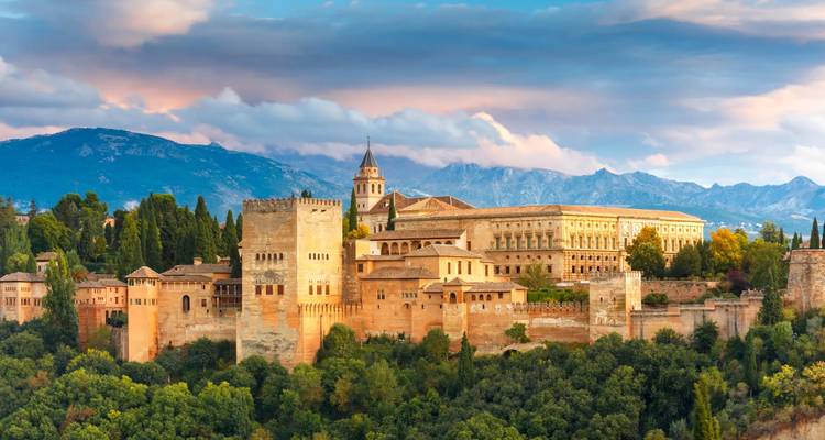 Spain’s Alhambra palace complex crowns a wooded hill with snow-capped peaks behind at dusk.