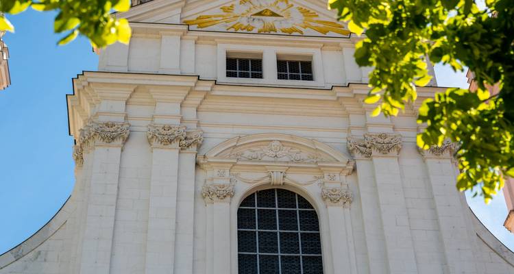 Detail einer weißen barocken Kirchenfassade, umrahmt von leuchtend grünen Blättern vor einem klaren Himmel.