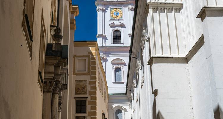 Eine enge Gasse gibt den Blick auf einen Glockenturm und klassische Säulen unter strahlend blauem Himmel frei.