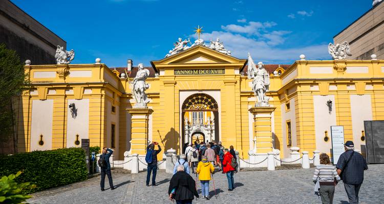 Besucher betreten ein leuchtend gelbes barockes Tor, das mit Statuen geschmückt ist, auf einem historischen Klosterhof.