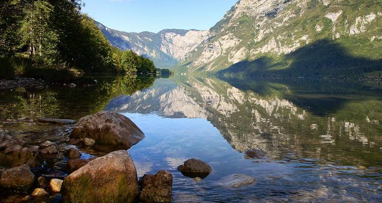 Lac alpin cristallin reflétant parfaitement les pentes boisées et les montagnes escarpées sous un ciel bleu.