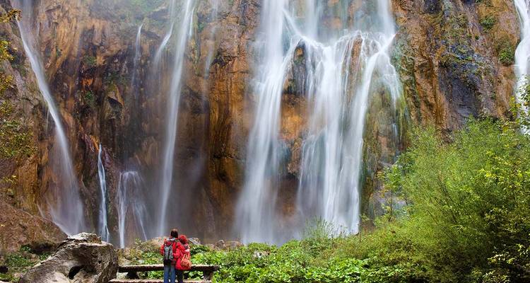 Deux routards regardent vers le haut de multiples chutes d'eau en cascade qui se déversent le long d'une falaise escarpée dans un environnement luxuriant.