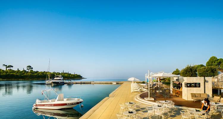 Terrasse de café en bord de mer avec des yachts amarrés et une mer bleue calme sous un ciel matinal lumineux.