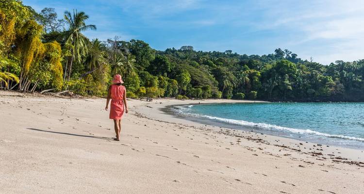 Woman in a pink dress strolls along a secluded tropical beach backed by jungle.