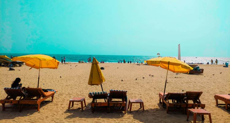 Des rangées de transats en bois et de parasols jaunes bordent une large plage de sable face à une mer scintillante.