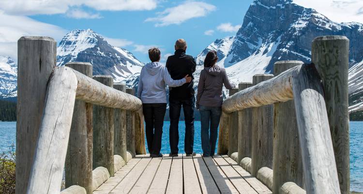 Trois voyageurs se tiennent debout sur un quai en bois face à un lac glaciaire et à de majestueuses montagnes enneigées dans les Rocheuses canadiennes.