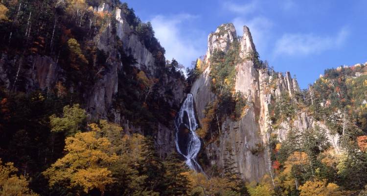 Ein hohes Wassergefall stürzt über eine dramatische Felsklippe herab, umgeben von farbenfrohem Herbstwald.