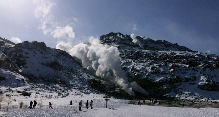 Rauchfahnen steigen von einem schneebedeckten vulkanischen Berghang auf, während Besucher die winterliche Landschaft erkunden.