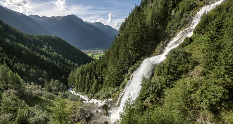 Hoge waterval die neerstroomt in een groen alpendalletje omringd door dicht sparrenbos