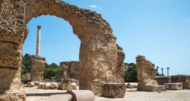 El arco de arenisca desgastada enmarca las columnas y bloques restantes de las antiguas ruinas romanas de Cartago contra un cielo despejado.