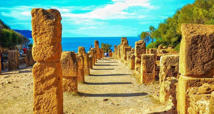 Las columnas de piedra romana iluminadas por el sol bordean una antigua avenida junto al mar con el mar azul brillante en la distancia.