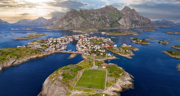 Vue aérienne spectaculaire de Henningsvær avec son terrain de football sur une minuscule péninsule entourée par la mer bleue et des îles.