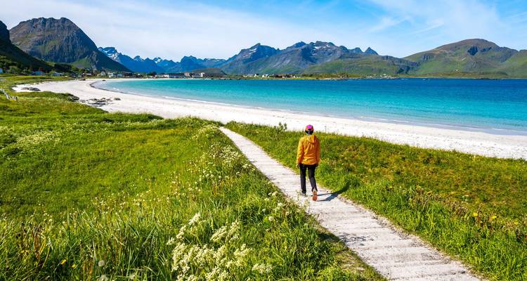 Un randonneur suit une passerelle en bois vers une plage arctique à sable blanc et une mer turquoise dominées par des pics dentelés.