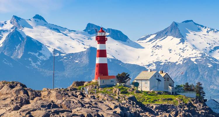 Le phare rouge et blanc se dresse sur un îlot rocheux face à des montagnes enneigées spectaculaires sous des ciels bleu clair.
