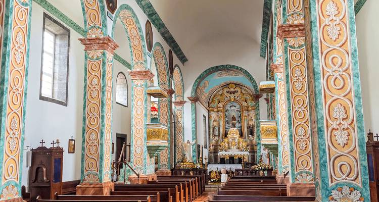 Ornately painted church interior with colorful patterned columns, arched windows and wooden pews leading to a golden altar.