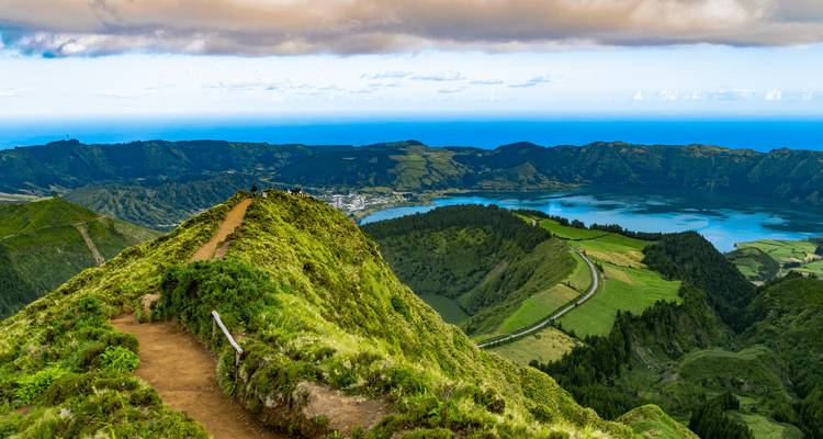 Ridgeline hiking trail leading toward emerald lakes inside a vast volcanic caldera with the Atlantic in the distance.