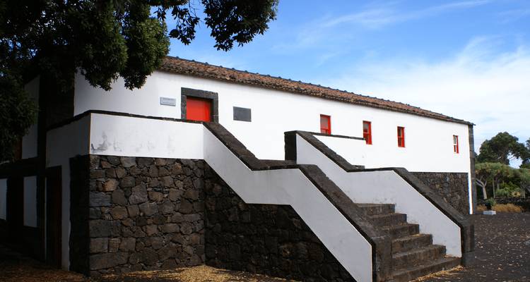 Simple white building with red doors and volcanic stone foundation under a bright blue sky.