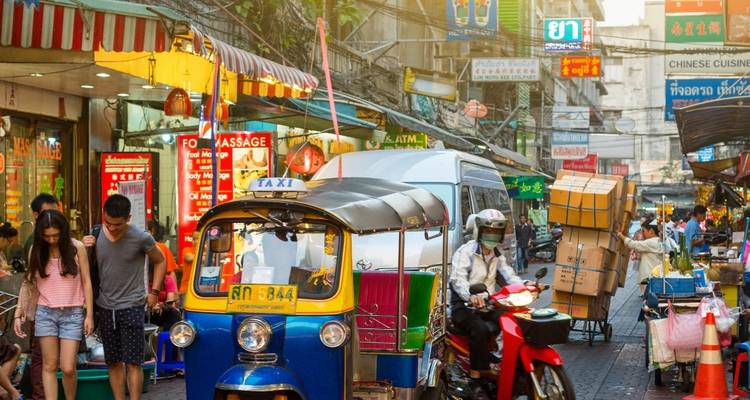 Scène animée d'une rue de Bangkok avec un tuk-tuk bleu et jaune, des piétons, des panneaux de signalisation et des façades de boutiques colorées en plein jour.
