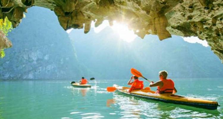 Les voyageurs vêtus de gilets de sauvetage orange naviguent en kayak dans une eau émeraude sous une ouverture de grotte en calcaire dramatique avec la lumière du soleil qui s'écoule à l'intérieur.