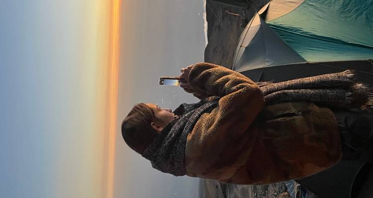Ingepakt in warme lagen fotografeert een trekker een gloeiendo horizon vanaf naast opgezette tenten op de berg.