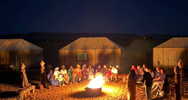 Reizigers verzamelen zich rond een flink brandend kampvuur tussen woestijntenten onder een heldere nachtelijke hemel.