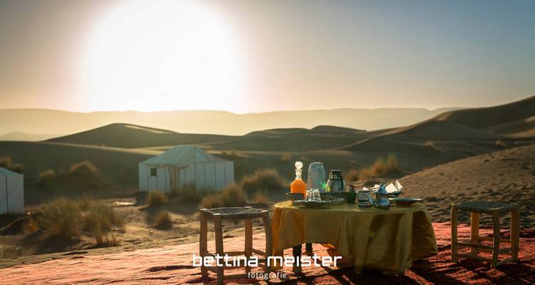 Ontbijttafel op woestijnzand bij zonsopgang met tenten en golvende duinen op de achtergrond.